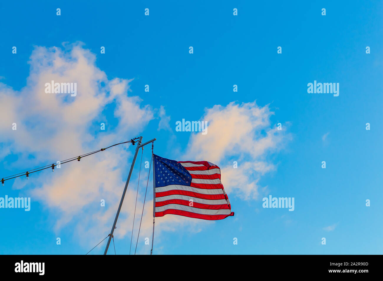 An American ensign blows in the wind as it hangs from a gaff at the stern of a boat. The flag flies against a blue sky with white clouds. Stock Photo