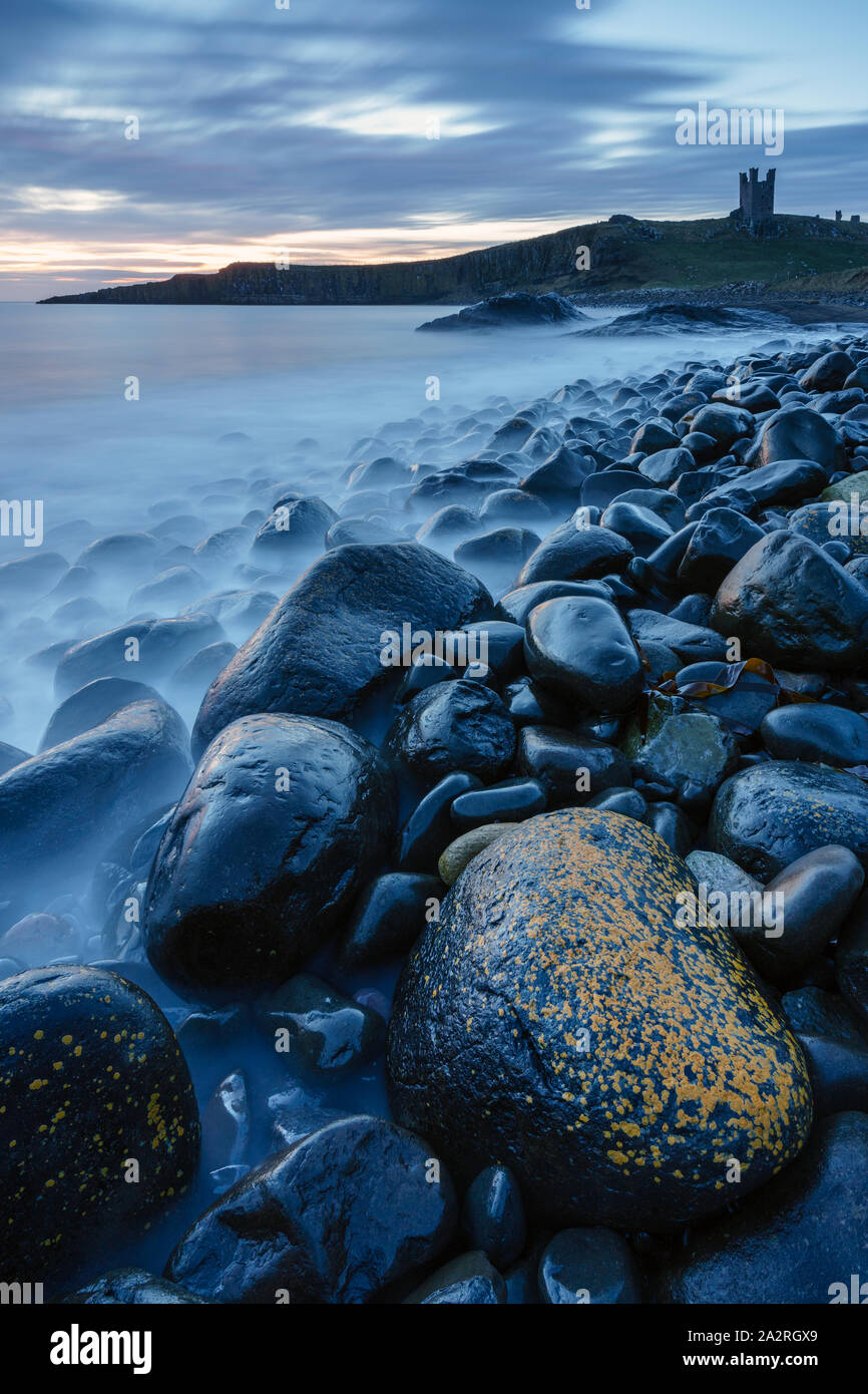 The imposing ruins of Dunstanburgh Castle tower above the dolerite boulder strewn Embleton Bay as dawn light begins to highlight the scene. Stock Photo