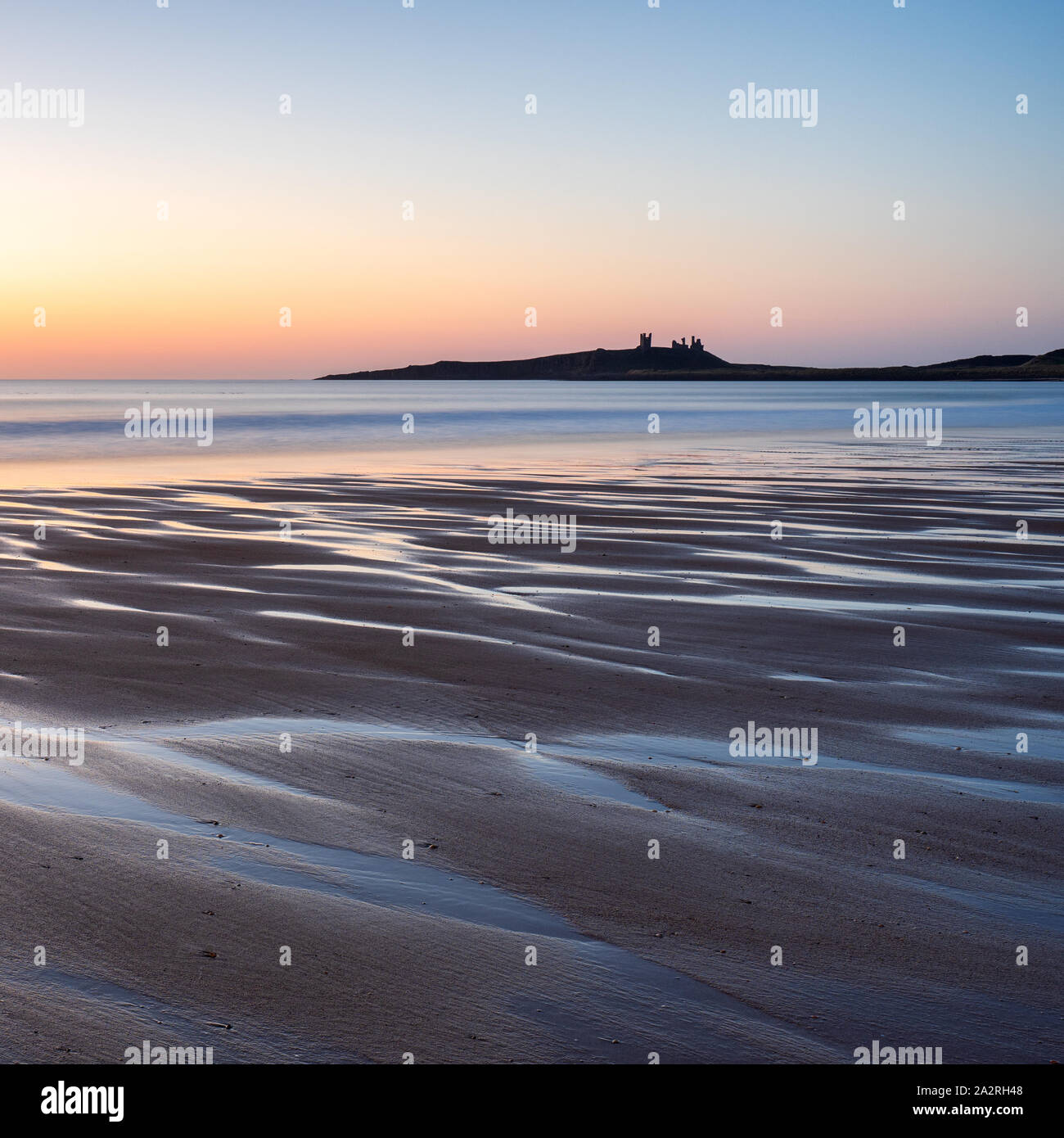 Beautiful patterns in the sand at Embleton Bay reflect the pre-sunrise sky with the imposing ruins of Dunstanburgh Castle on the horizon. Stock Photo