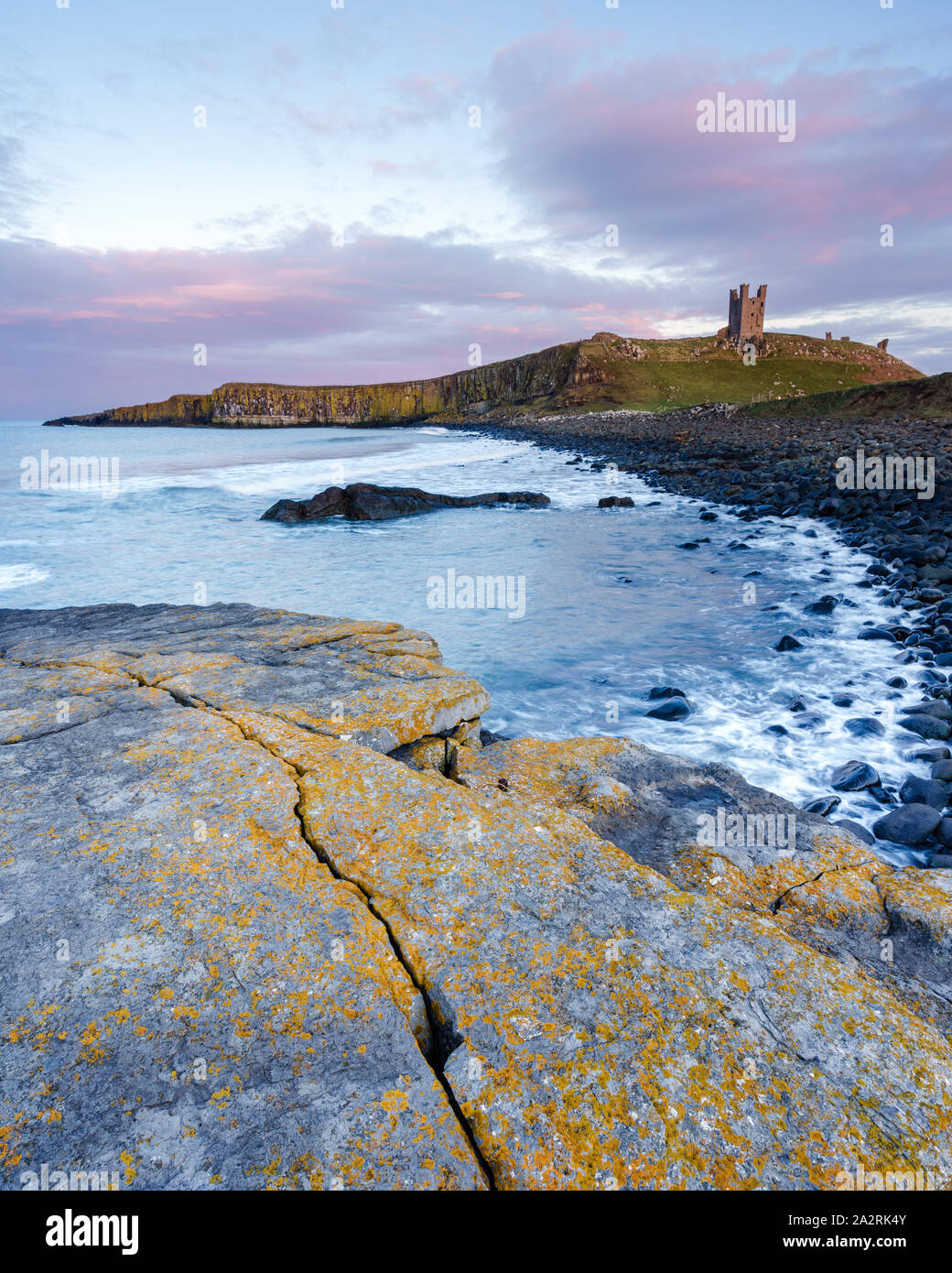 Sunset light illuminates Dunstanburgh Castle above Embleton Bay with the vibrant colours and textures of the rocky coastline filling the foreground. Stock Photo