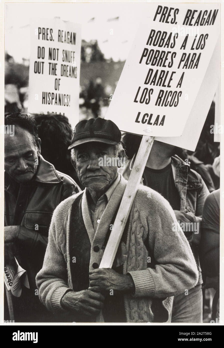 Reagan la roba a los pobres Migrant farm worker at a demonstration, Washington, D.C. / / Espada.; Photograph shows an elderly man in the Solidarity Day march, holding a picket sign with the message: Pres. Reagan robas a los pobres para darle a los ricos, LCLAA, in a crowd at a protest. A sign in the background has the message: Pres. Reagan, do not snuff out the dreams of Hispanics!; Stock Photo
