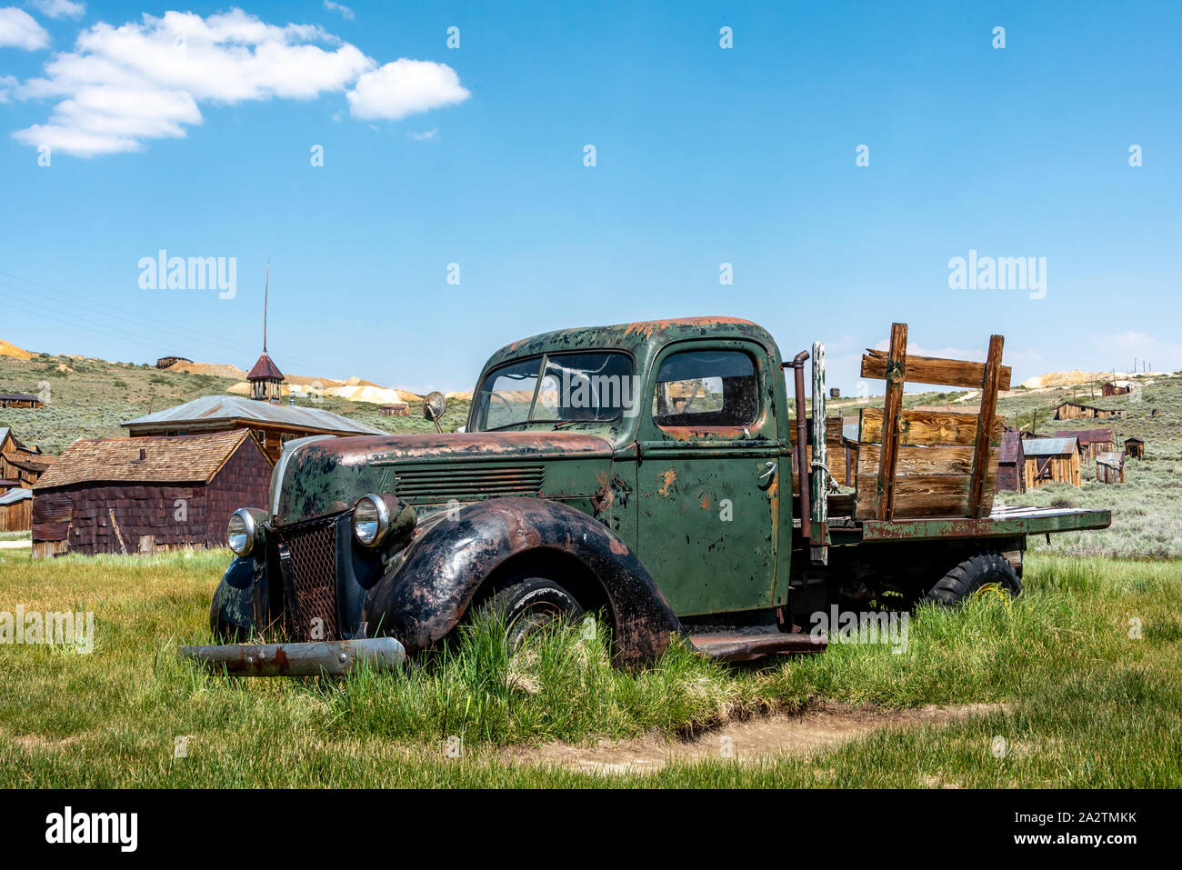 An old green pickup truck with flat bed and wooden rails deteriorates where abandoned at Bodie State Historic Park and ghost town. Grass grows up arou Stock Photo