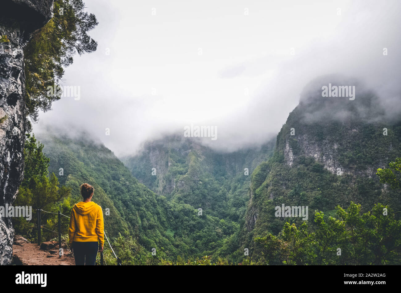Young woman in yellow sweatshirt on a viewpoint in Levada Caldeirao Verde, Madeira, Portugal. Green scenic mountains in fog, misty landscape. Female traveler. Instagram, hipster filter. Stock Photo