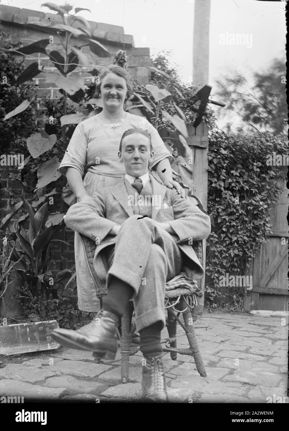 Glass Negative - Couple in Garden, circa 1920s - 1930s, Black and white 1/6th plate glass negative of a couple in a back garden or courtyard. The man is seated cross legged in a chair and the woman stands behind him. collection of products, promotional materials, photographs and working life artefacts, when the Melbourne manufacturing plant at Coburg closed down Stock Photo