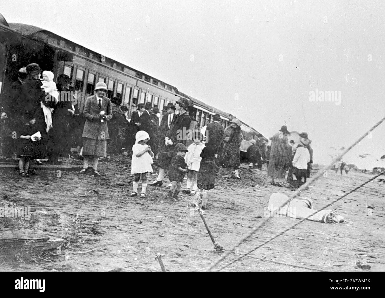 Negative - Group Inspecting 'Better Farming Train', Underbool, Victoria, 1927, A group, predominantly female, beside railway carriages. This was the 'Better Farming Train'. The women a wearing skirts and jackets, and dress shoes. Some of the women are wearing hats. A group of children are standing in front of the women, the girls are wearing short dresses and long socks, and the boys are wearing shorts and jackets. On the left, a woman is holding a baby which is wrapped Stock Photo