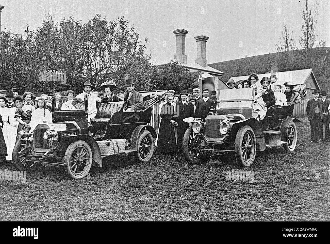 Negative - Wedding Party in Cars Surrounded by Onlookers, Scrubhill Dean, Victoria, 1909, The first time cars were used in Dean for a wedding. The wedding party (in the cars) is surrounded by onlookers. The building behind the group has two chimneys and a water tank Stock Photo