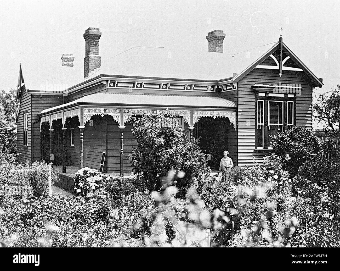 Negative - Woman by Verandah, Bullarook District, Victoria, 1927, A woman stands by the verandah of a house. The house appears well kept and newly painted (?). The garden in front of the house is in full bloom. There are three visible chimneys on the house and wrought iron lace work on the verandah Stock Photo