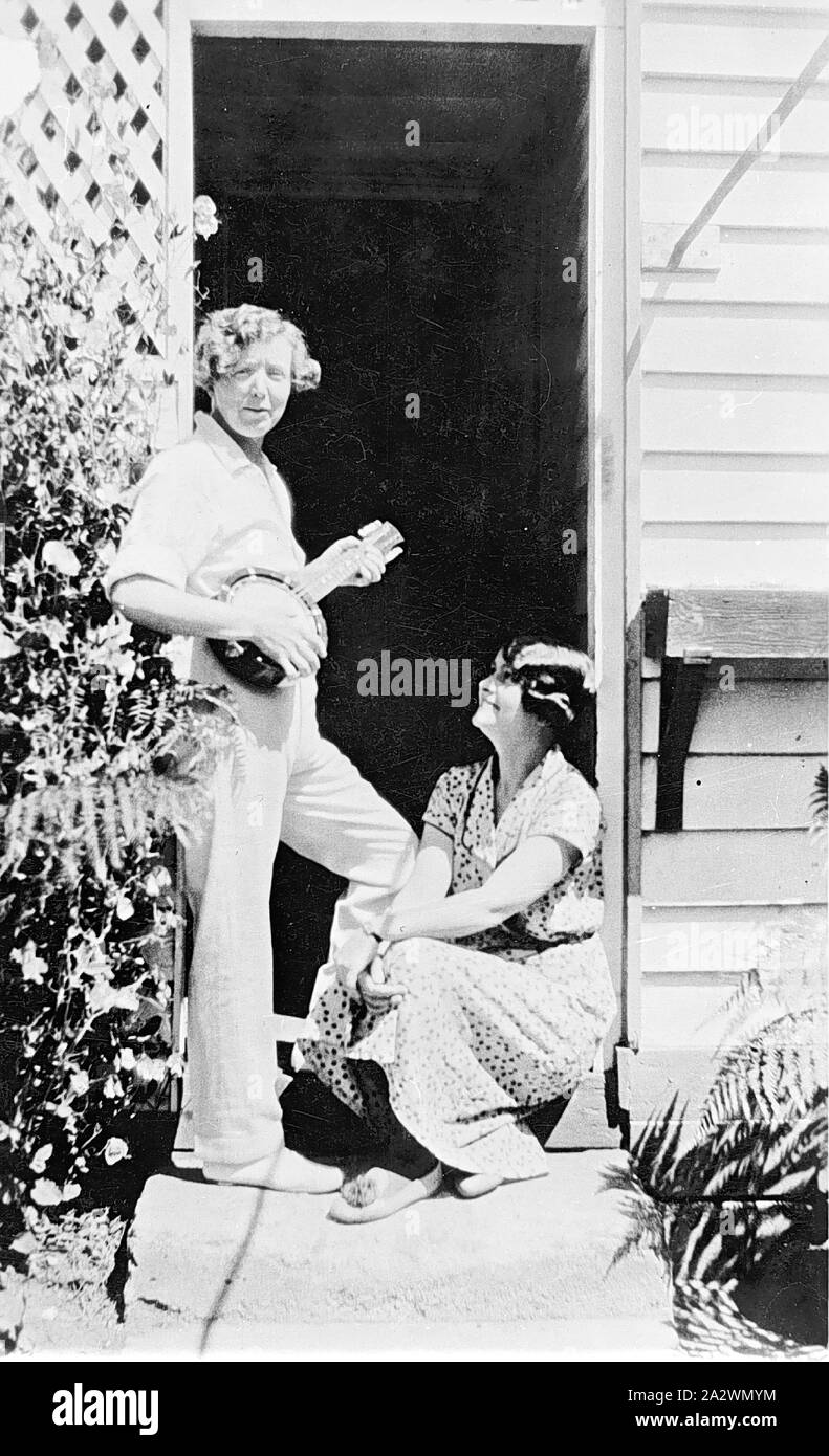 Negative - Woman Playing a Banjo, Ararat, Victoria, circa 1925, A woman playing a banjo. She is wearing slacks and standing in a doorway. Another woman is sitting on the step below looking up at her Stock Photo