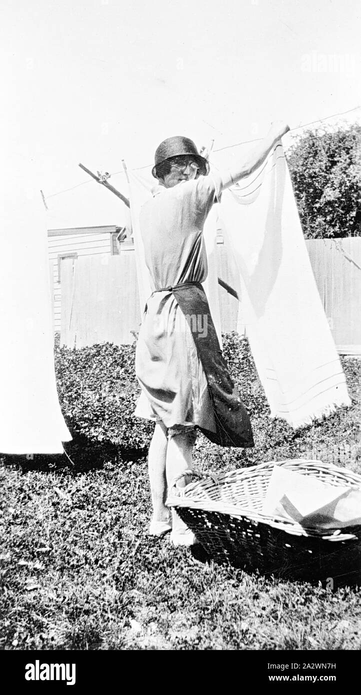 Negative - Woman Hanging Out Clothes, Western District (?), Victoria, circa 1935, A woman hanging out clothes. She wears a hat and an apron containing pegs in the front pocket. A washing basket rests at her feet. There is a clothes prop in the background Stock Photo