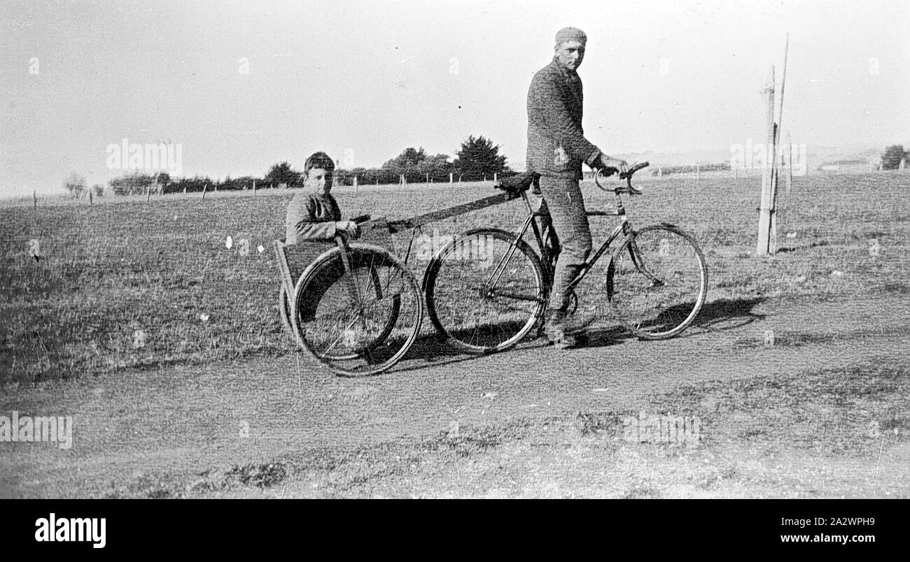 Negative - Larpent, via Colac, Victoria, circa 1920, Young man riding a bicycle with upturned handle bars. The bicycle is hitched to a home-made wooden cart mounted on two bicycle wheels. A boy sits in the cart. The young man wears long pants and jacket with a tight fitting cap or knotted handkerchief on his head. They are riding along a dirt track with fencing and trees visible in the background Stock Photo