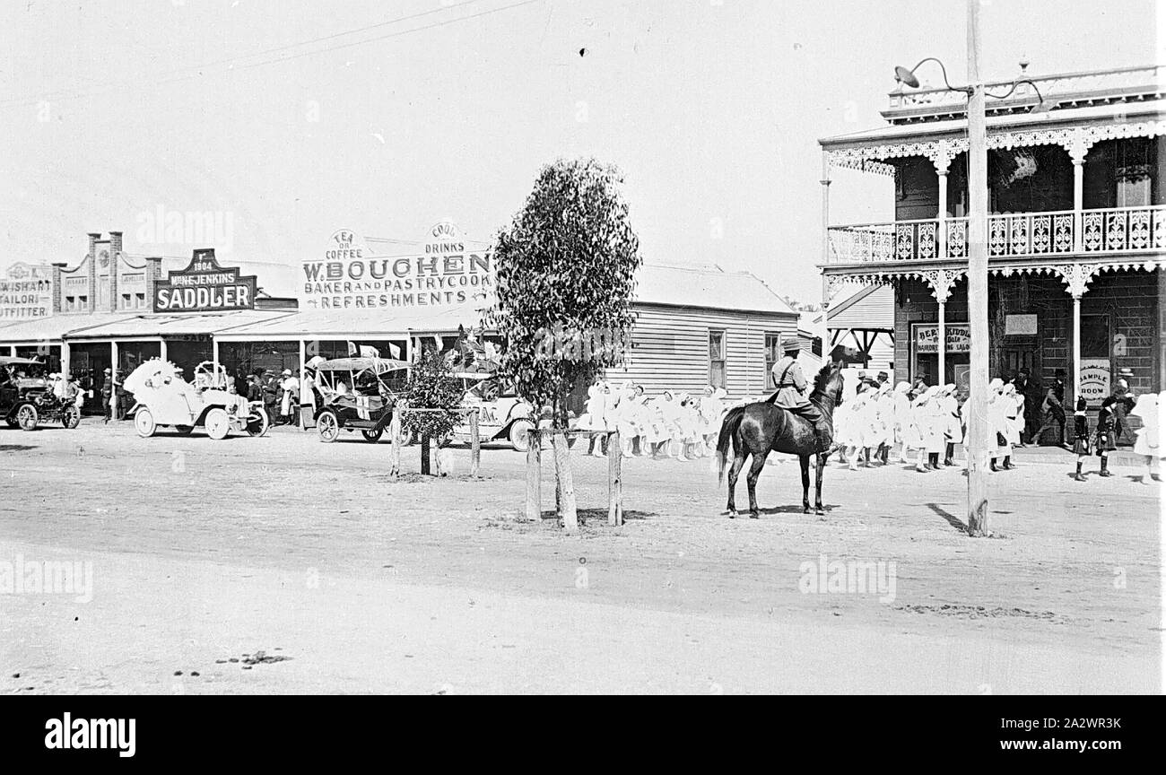 Negative - Rainbow, Victoria, circa 1925, The main street in rainbow. There is a parade of children followed by a number of motor cars. A soldier on horseback watches the parade. A saddler's, a pastrycook and a hotel are in the background Stock Photo