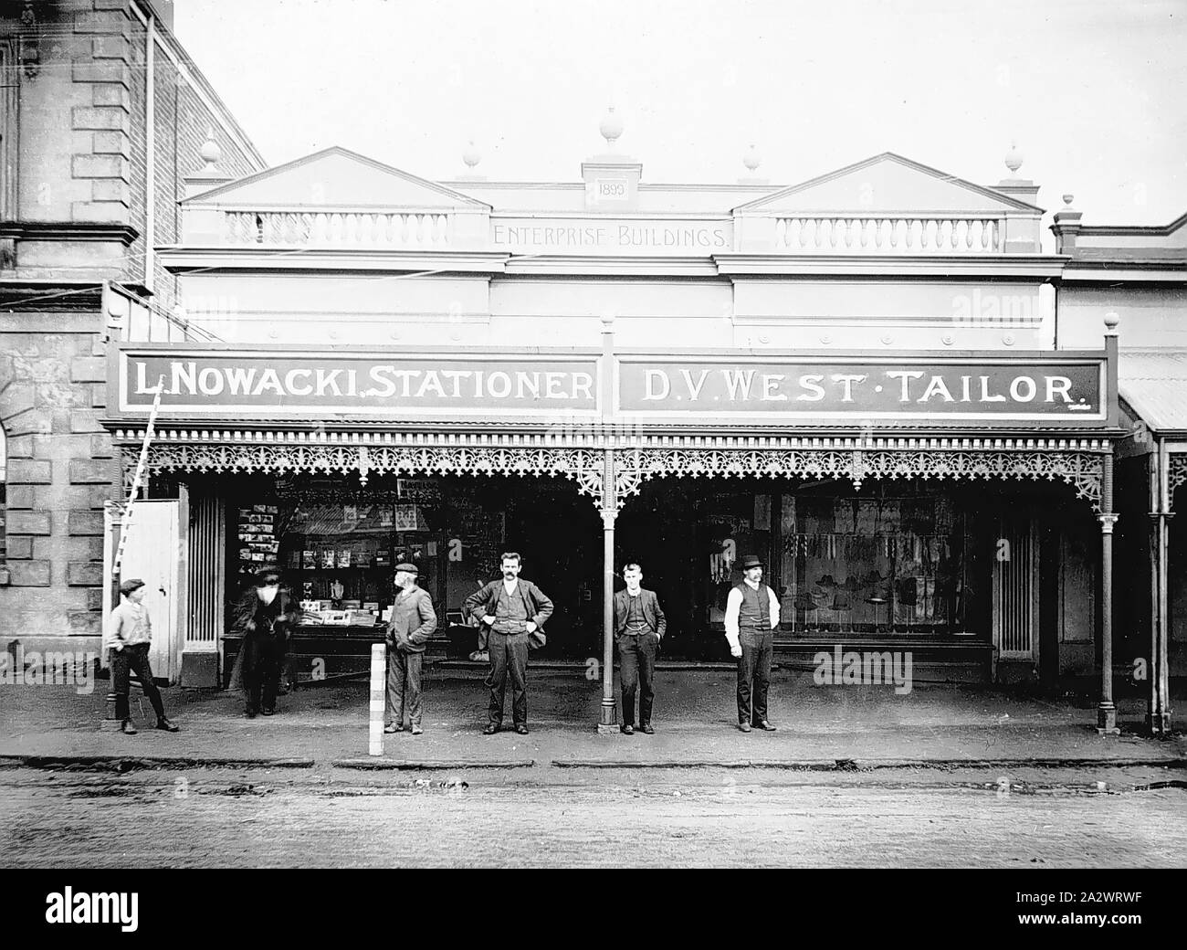 Negative - Casterton, Victoria, 1902, A group of men outside two shops forming the Enterprize Building, constructed in 1899. The two shops are 'L. Nowacki, stationer' and'D. V. West Tailor'. The old Mechanics Institute is on the left. There is wrought iron lacework above the verandah of the shops Stock Photo