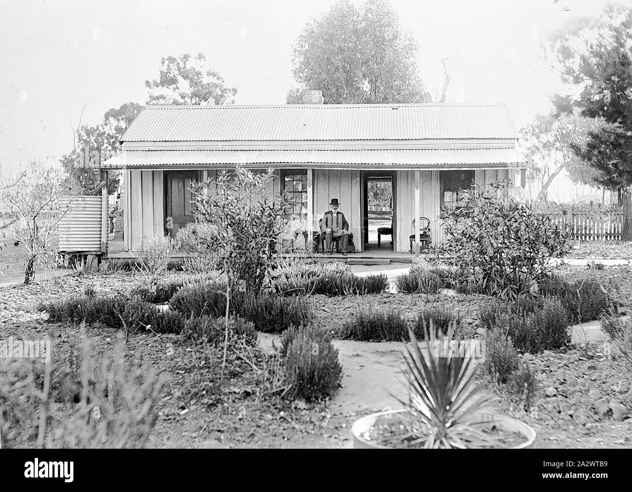 Negative - Bundalong, Victoria, 1907, A man seated on the verandah of a small house. The front door is open and the backyard can be seen through an open back door. There is a garden in front of the house and a water tank at the side Stock Photo