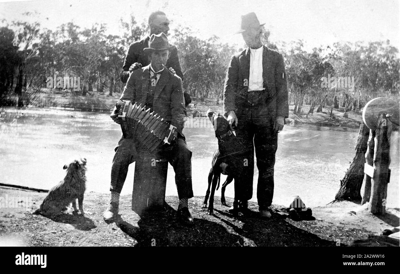 Negative - Mildura District, Victoria, circa 1925, Three men and two dogs on the bank of the Murray River. One man is playing an accordion while a second man stands behind him with his hands on his shoulders. The third man restrains one of the dogs. There is a grindstone on the right Stock Photo