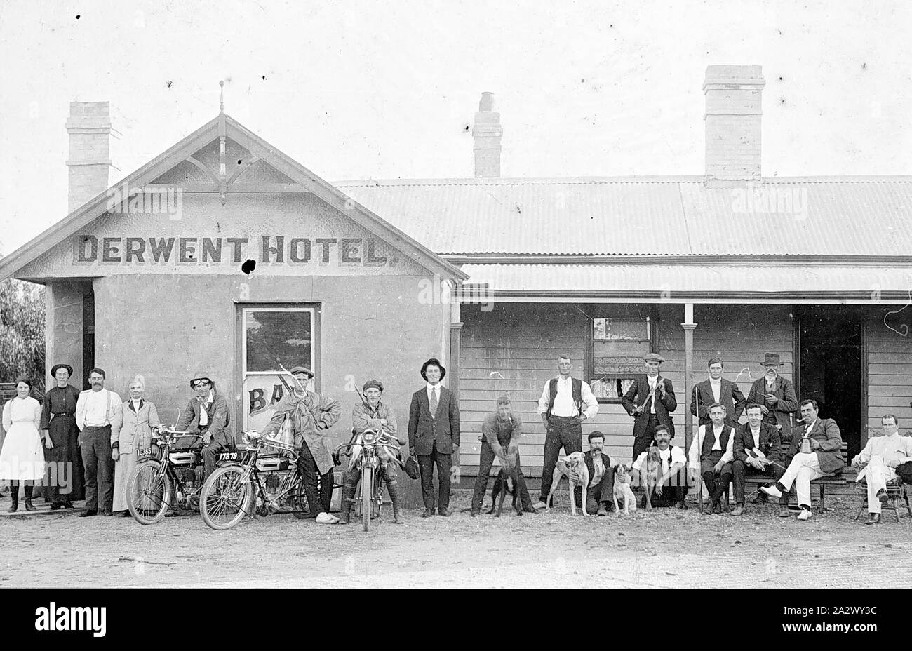 Negative - Motor Cycle Hunting Party with Group of Locals Outside Derwent Hotel, Batesford, Victoria, circa 1915, A group of eighteen (18) people standing and seated in front of the Derwent Hotel at Batesford. Three of the group are standing beside or astride early motor bikes, identified as being pre-World War I model Triumph motor cycles of single cylinder belt-driven type, with acetylene headlamps. The men are wearing hunting outfits and have motor cycle googles, and have rifles slung over their shoulders Stock Photo