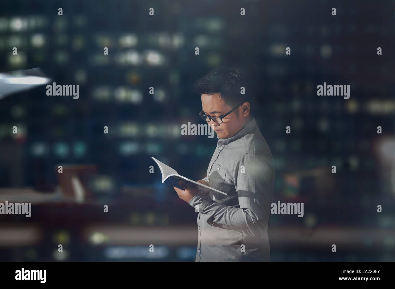 A side view shot of a young man in long sleeves and glasses peacefully reading a book inside a dark office room at night time. Stock Photo