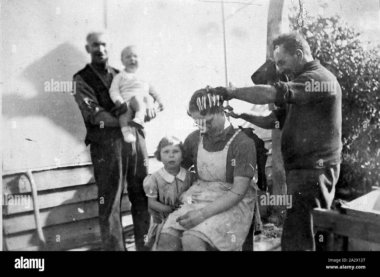 Negative - Gunbower District, Victoria, 1939, A man curling a woman's hair. A small girl stands beside her and a man holds a crying baby in the background Stock Photo