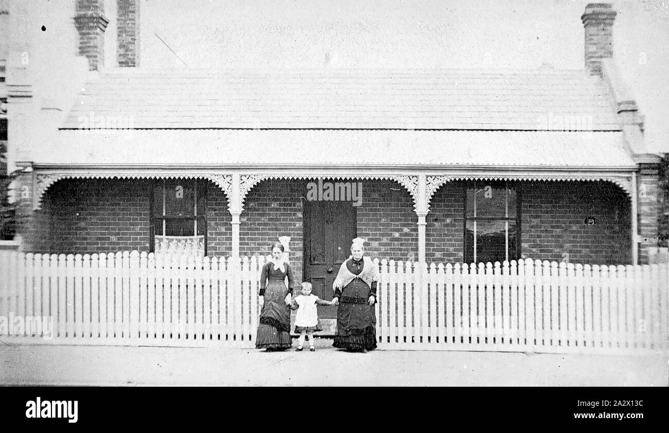 Negative - North Melbourne, Victoria, circa 1885, Two women and a small child stand in front of their brick house. This house has a verandah decorated with iron lacework and brick chimneys. A picket fence runs along the front of their property Stock Photo