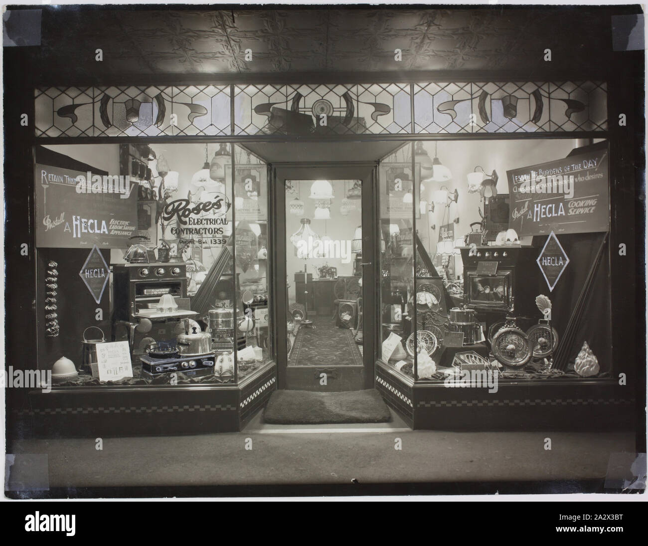 Photograph - Hecla Products in Roses Electrical Contractors, Brunswick, circa 1940, Black and white photograph of the shopfront of Roses Electrical Contractors, Sydney Road Brunswick. It features Hecla products in the display window including heaters [known then as electric fires], stoves, kettles and grillers. Several signs show Hecla slogans such as 'Lessen the burdens of the day' and 'Retain that youthful appearance'. Image features a double shop front window display of Hecla Stock Photo