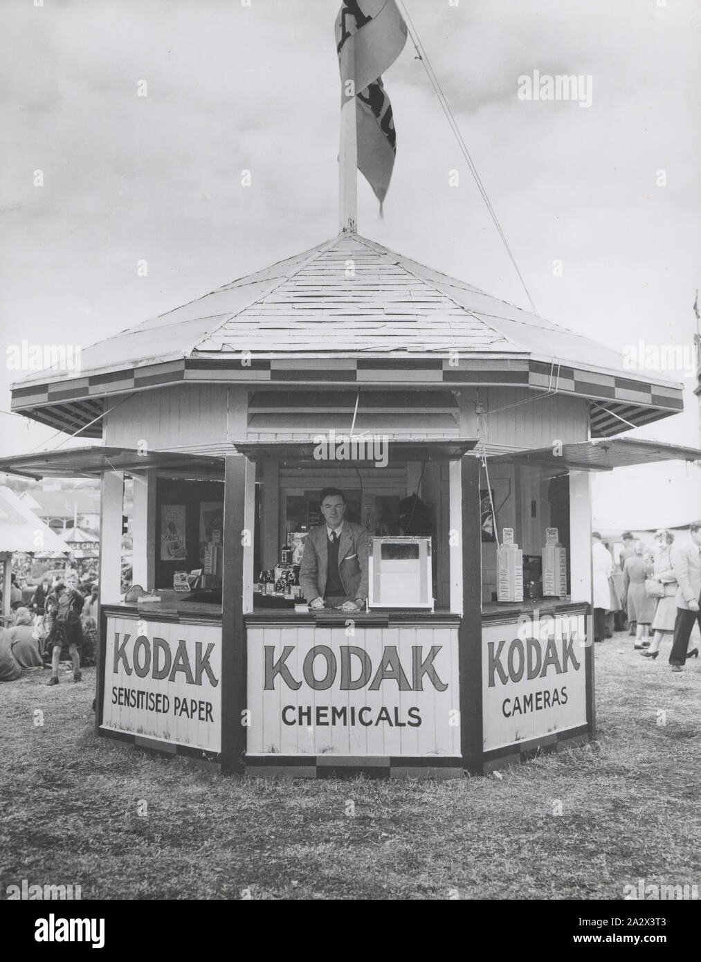 Photograph, Booth in Exhibition Grounds, circa 1930s, Black and white photograph of a Kodak Australasia Pty Ltd booth in an open-air exhibition ground, possibly the Royal Melbourne Show, or another interstate Royal Exhibition, circa 1930s. A Kodak staff member stands smiling to camera at the counter of the booth, and there are products arranged within. There is a large Kodak flag on top of the pointed roof and the windows of the booth are shuttered Stock Photo