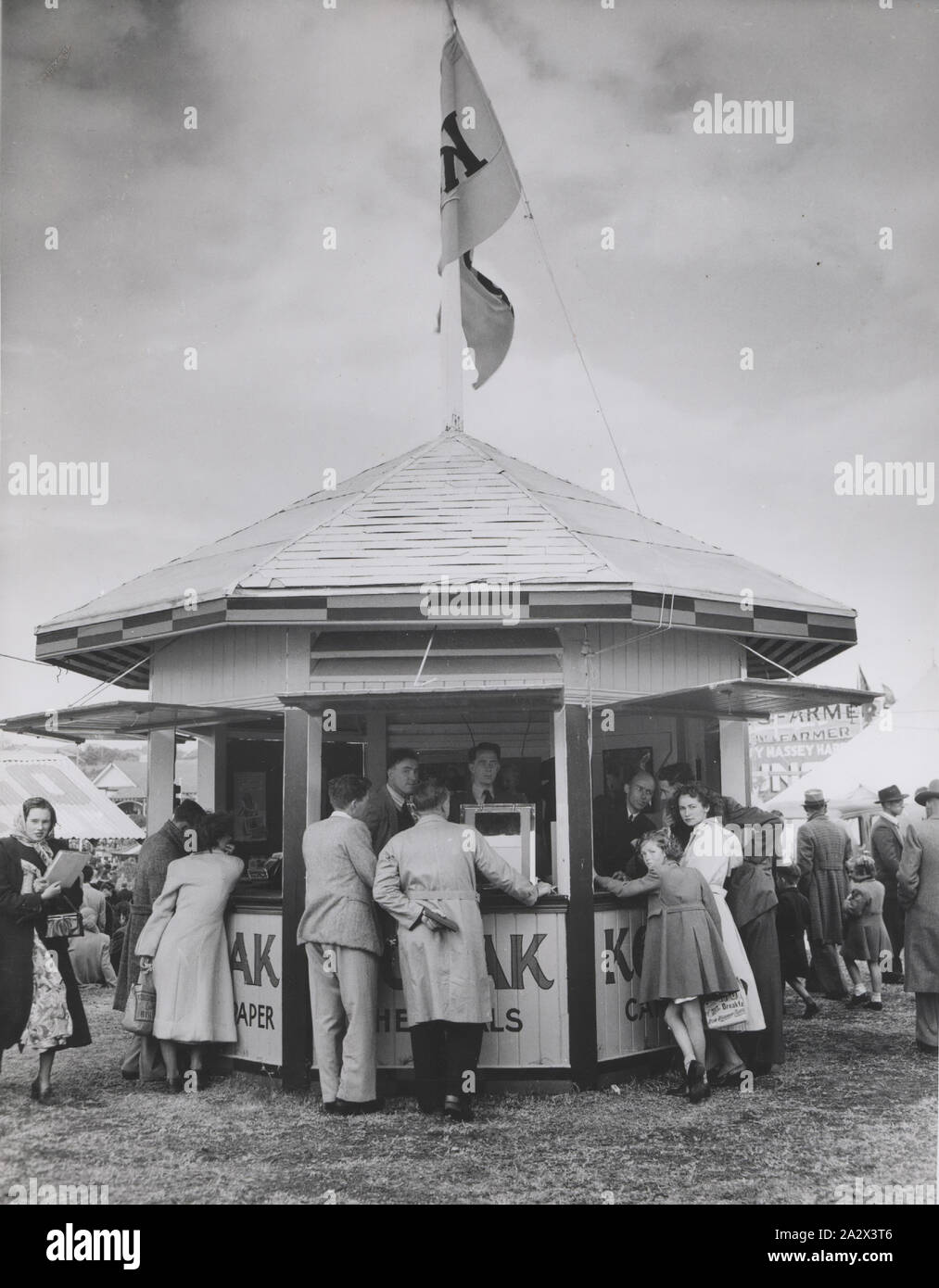 Photograph, Customers at Booth in Exhibition Grounds, circa 1930s, Black and white photograph of a Kodak Australasia Pty Ltd booth in an open-air exhibition ground, possibly the Royal Melbourne Show, or another interstate Royal Exhibition, circa 1930s. Six customers are lining up around the display and several male Kodak staff members are serving them, but many people have turned to look at the camera. This photograph is part of the Kodak collection of Stock Photo