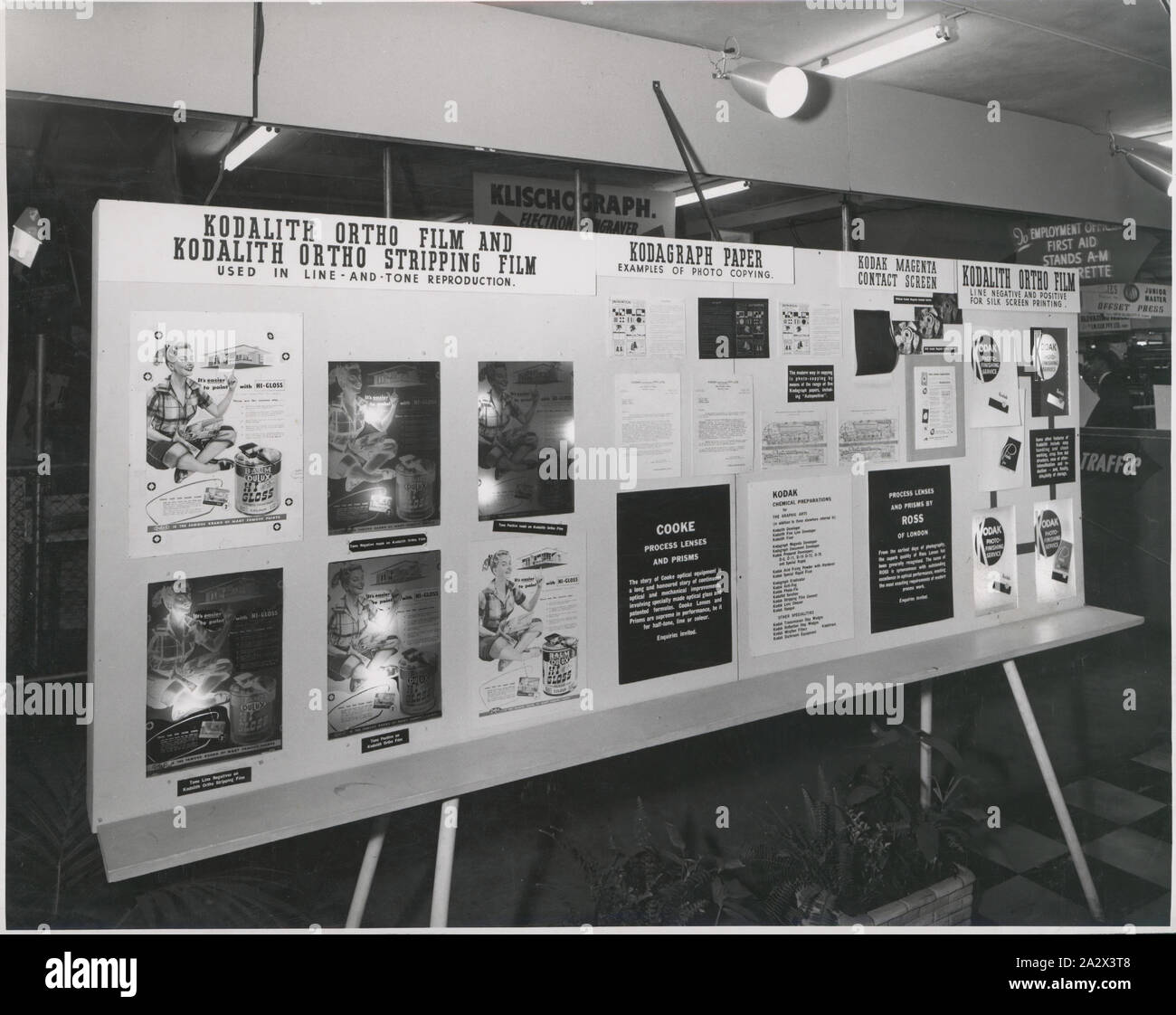 Photograph, Display Panel, Graphic Arts Products, Sydney, circa 1940s, Black and white photograph of a Kodak Australasia Pty Ltd exhibition display panel themed around graphic arts products, circa 1940s. Products include Kodalith Ortho films, Kodagraph Paper and Kodak Magenta Contact Screen. Beneath the panel, which is covered in posters and lit from above, is a chequerboard floor and decorative planter boxes with indoor plants. Other exhibition stands Stock Photo