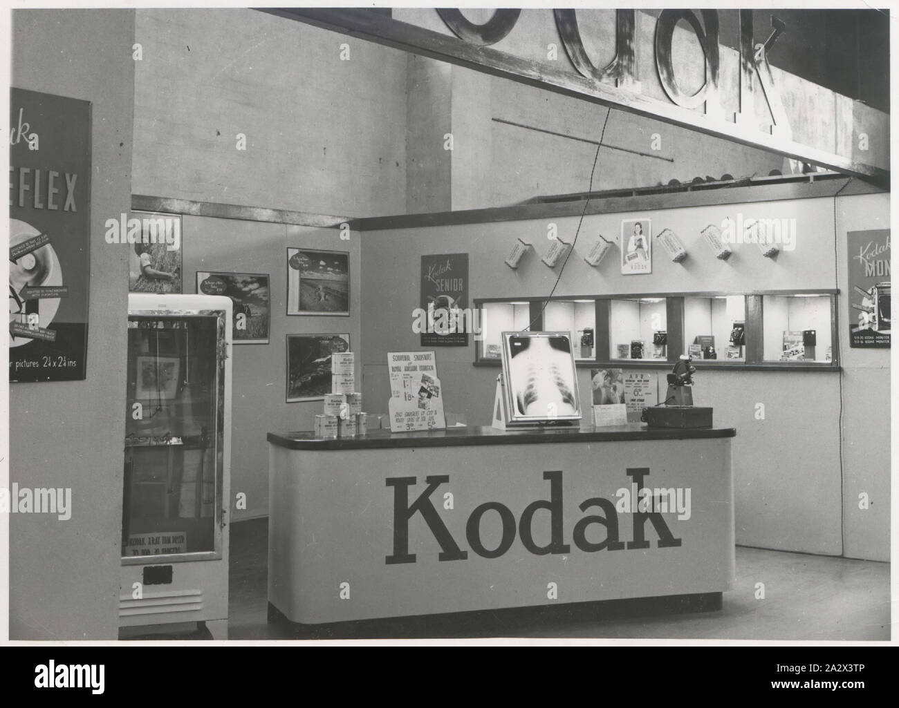 Photograph, Exhibition Stand, Chamber of Manufactures Exhibition, Adelaide, Mar - May 1947, Black and white photograph of a Kodak Australasia Pty Ltd exhibition stand or trade show booth at the Chamber of Manufactures Exhibition held from March to May 1947 in Adelaide. This view shows the service counter, drying cabinet, and wall of recessed display cameras with products and films arranged within. There are posters and x-ray film and photographs mounted around the display Stock Photo