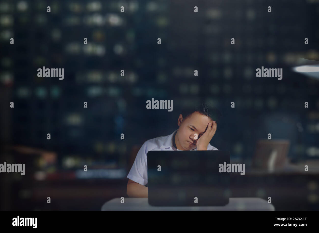 Young male student in school uniform feeling sleepy or almost falling asleep while studying and finishing a project using his laptop inside an office. Stock Photo