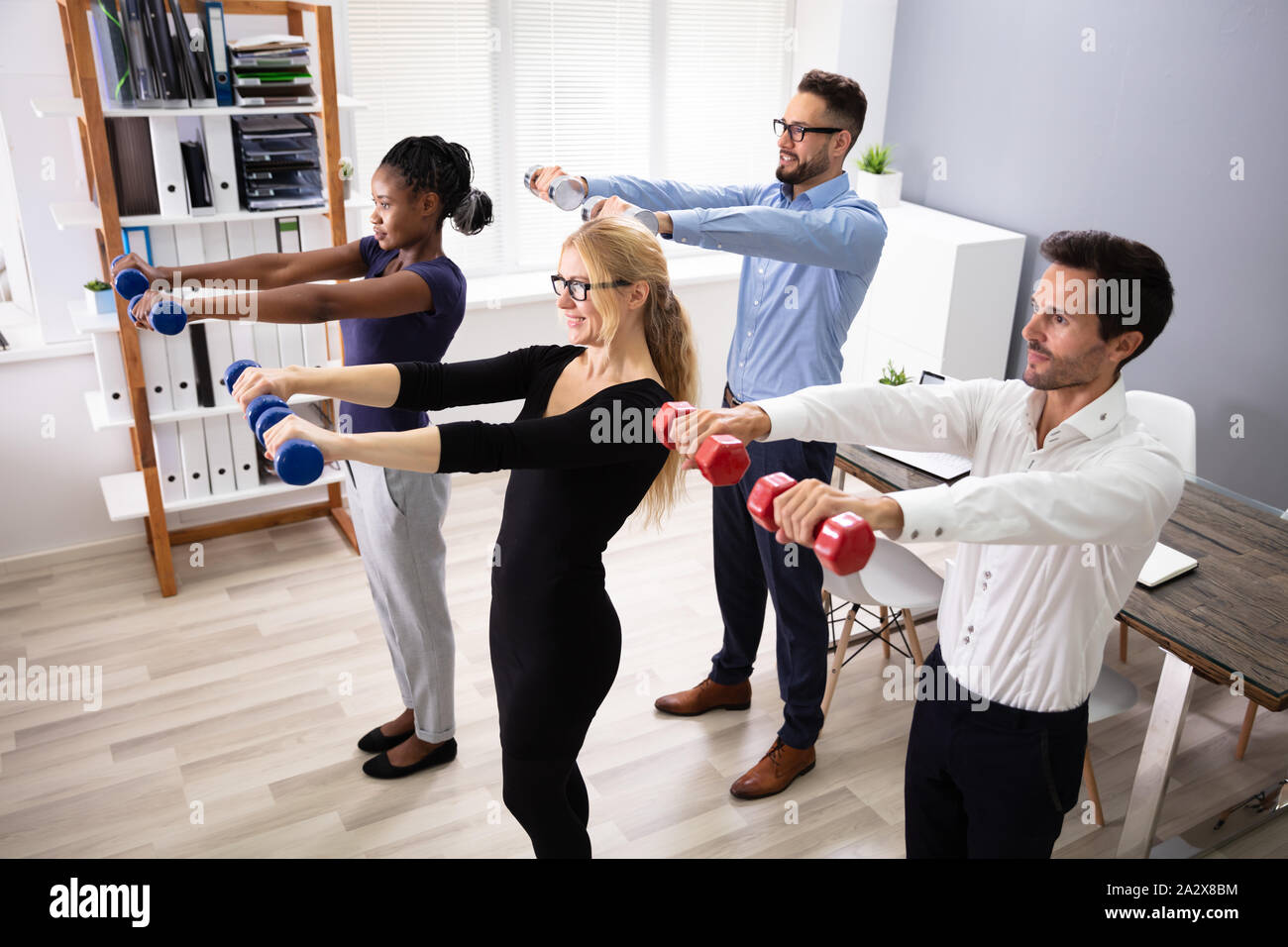 Happy Young Businesspeople Standing In Row Exercising With Dumbbells In Office Stock Photo