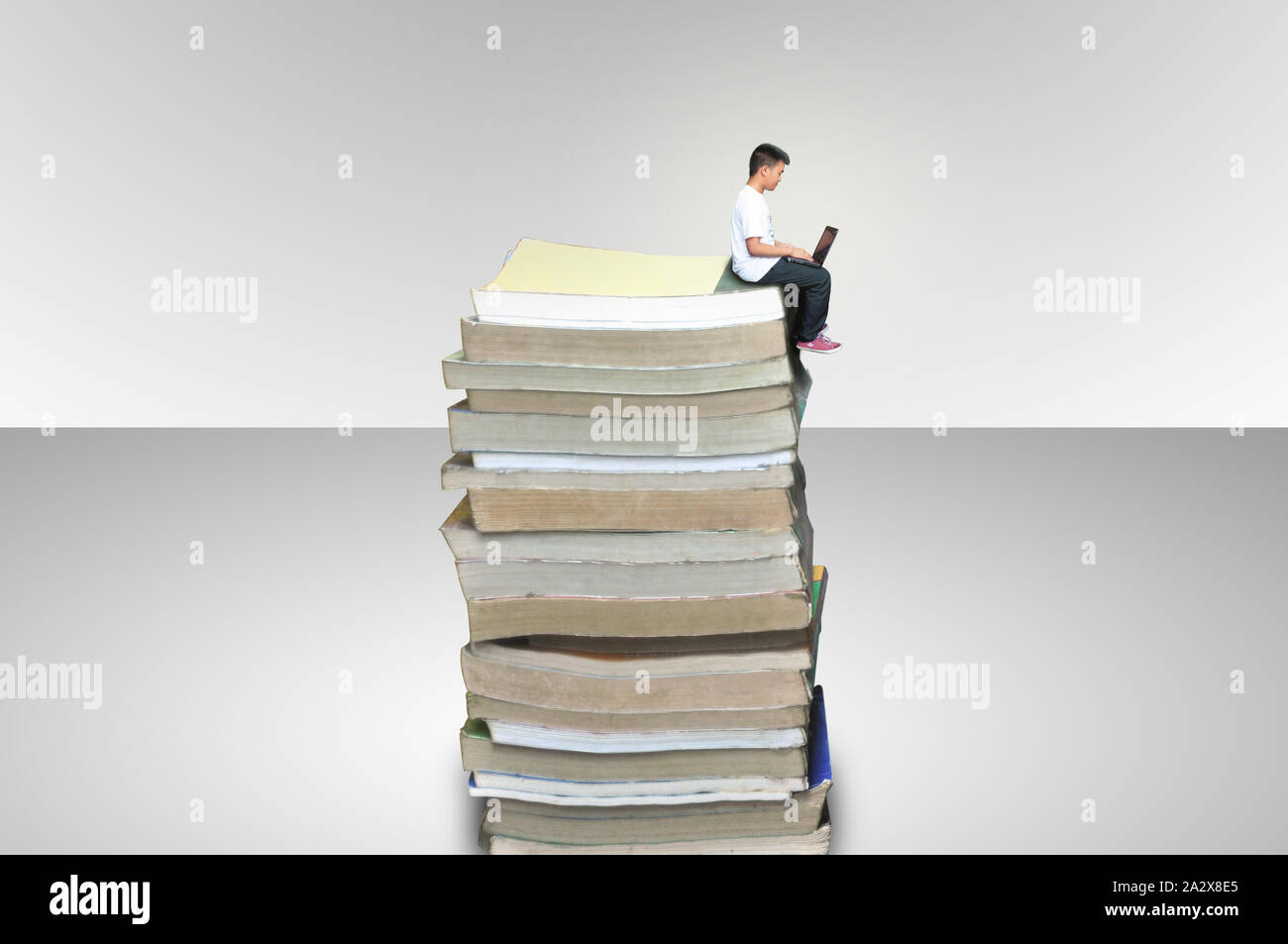 A boy sitting in a tower of books using his laptop in a white / gray background showing that the student had read many book and he is learning. Stock Photo