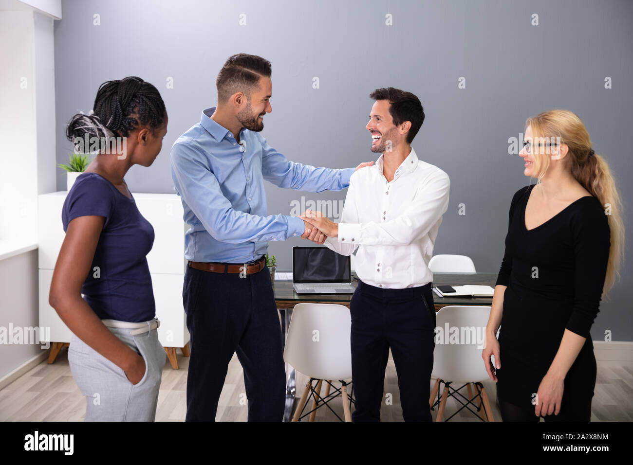 Women Looking At Happy Young Business Men Shaking Hands In Meeting Room Stock Photo