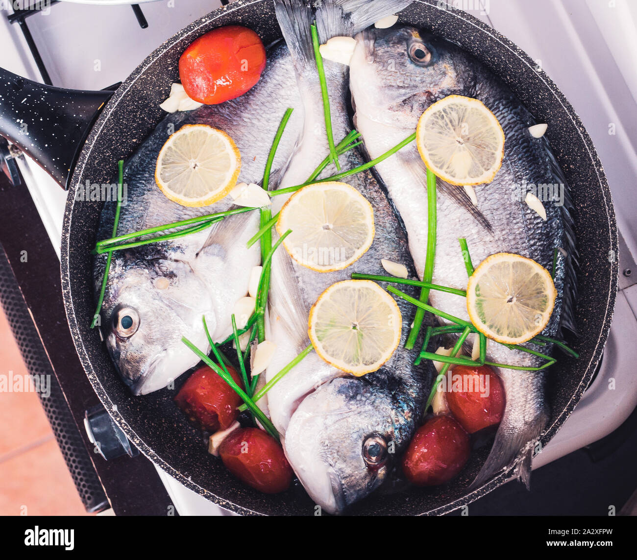 Raw orata fish on frying pan cooking with lemon, tomatoes and greenery. Top view Stock Photo