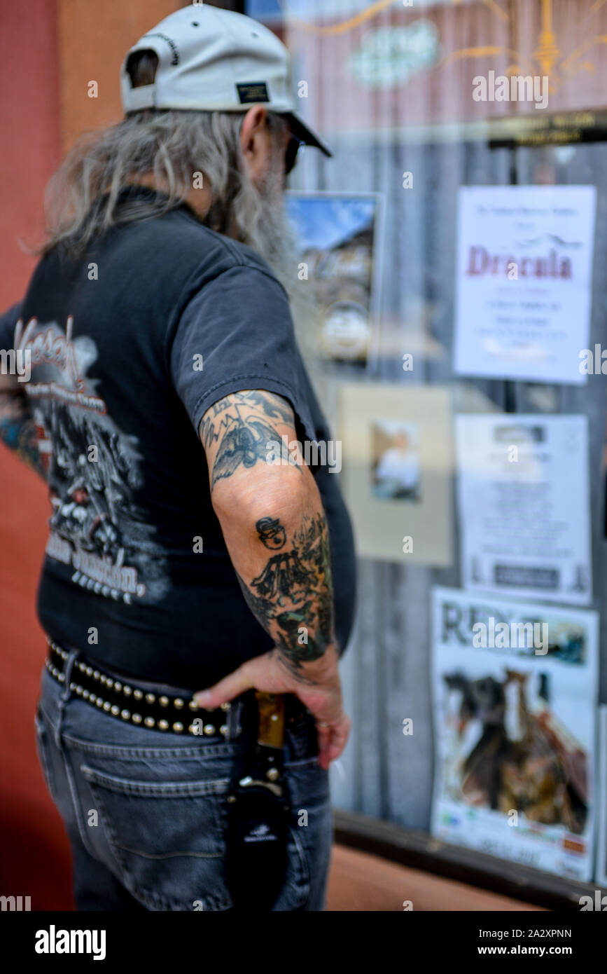 Rear view of long straggly haired middle aged man wearing baseball cap and knife on belt and arm tattoos studies storefront covered in posters Stock Photo