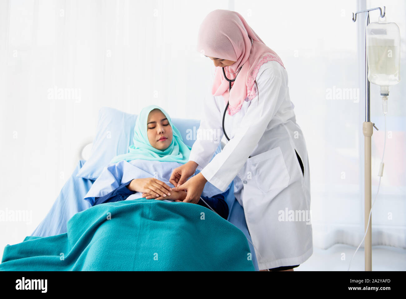 A beautiful young Muslim woman doctor is giving the saline solution to the patient in the arm who is lying on the bed in the hospital room. Stock Photo