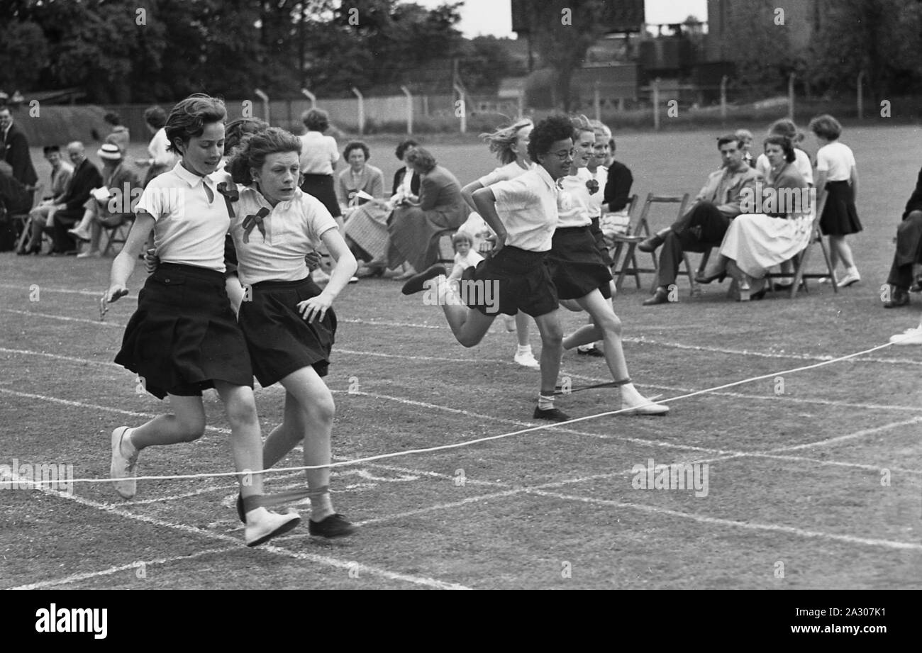 1950s, historical, primary school sports, schoolgirls taking part in a three-legged running race outside in a grass field, England, UK. A three-legged race is a fun running event involving pairs of children running with the left leg strapped to the right leg of another runner. Stock Photo