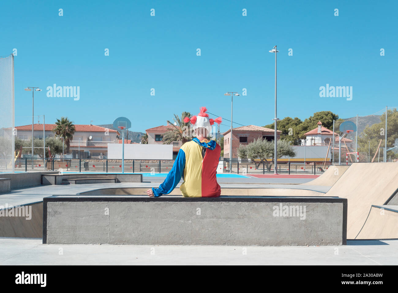 a scary clown, wearing a colorful yellow, red and blue costume, seen from behind, smoking a cigarette, sitting in an outdoor public sports complex Stock Photo