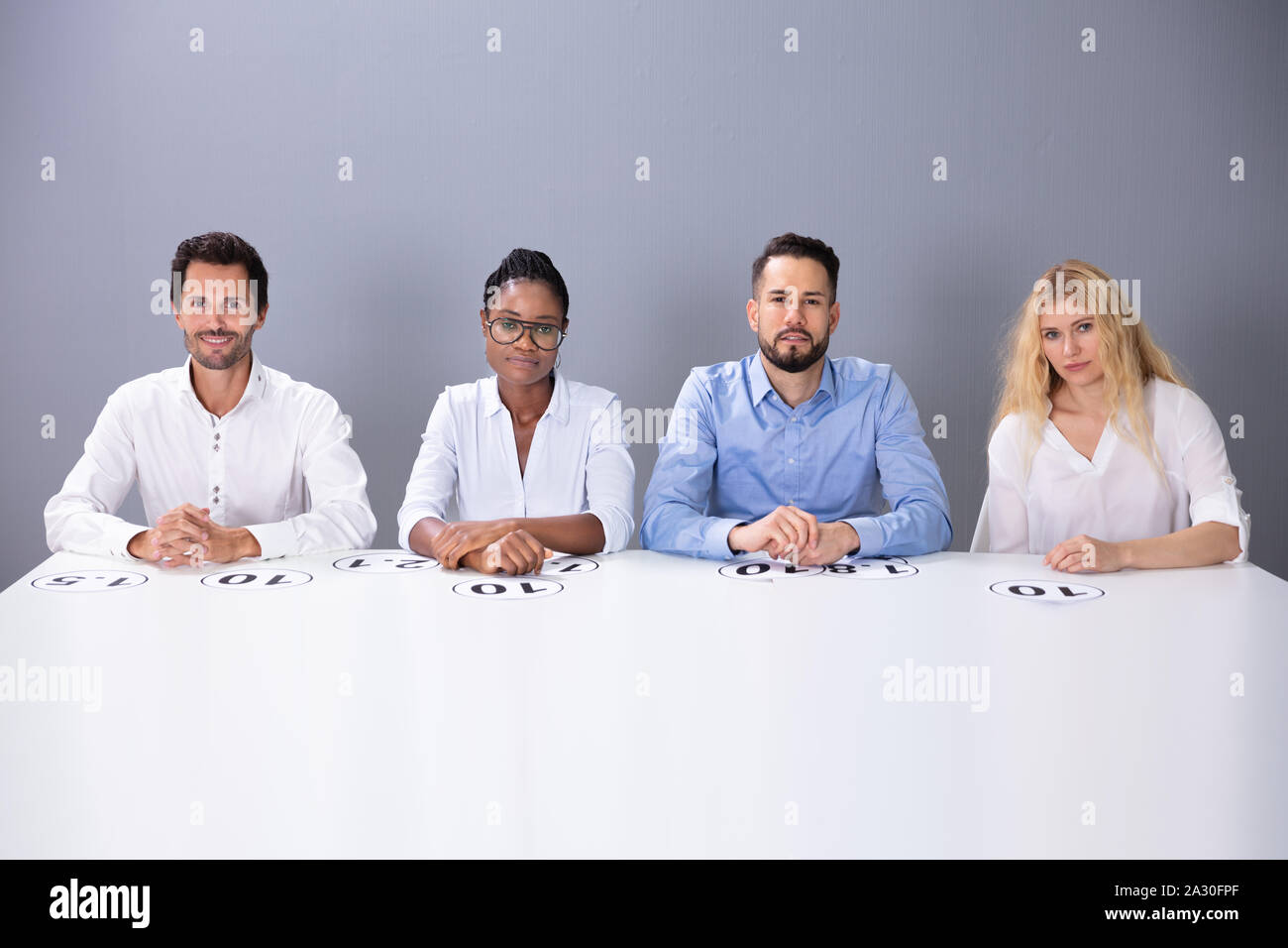 Multiracial Confident Judges Sitting On Chair With Scoring Points On Table Against Wall Stock Photo