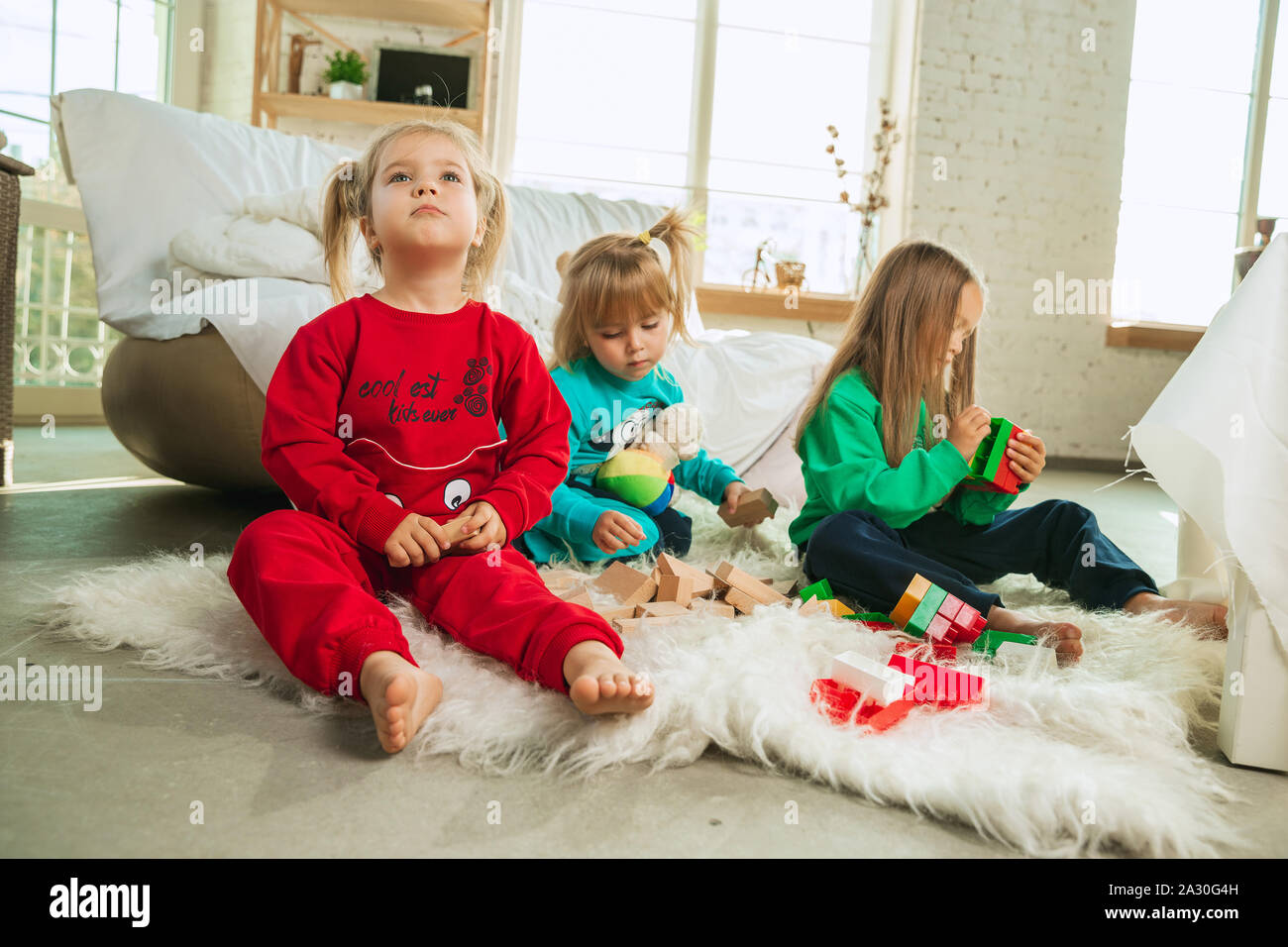 Little girls in soft warm pajamas playing at home. Caucasian children in colorful clothes having fun together. Childhood, home comfort, happiness. Sitting on the floor and making constructor. Stock Photo