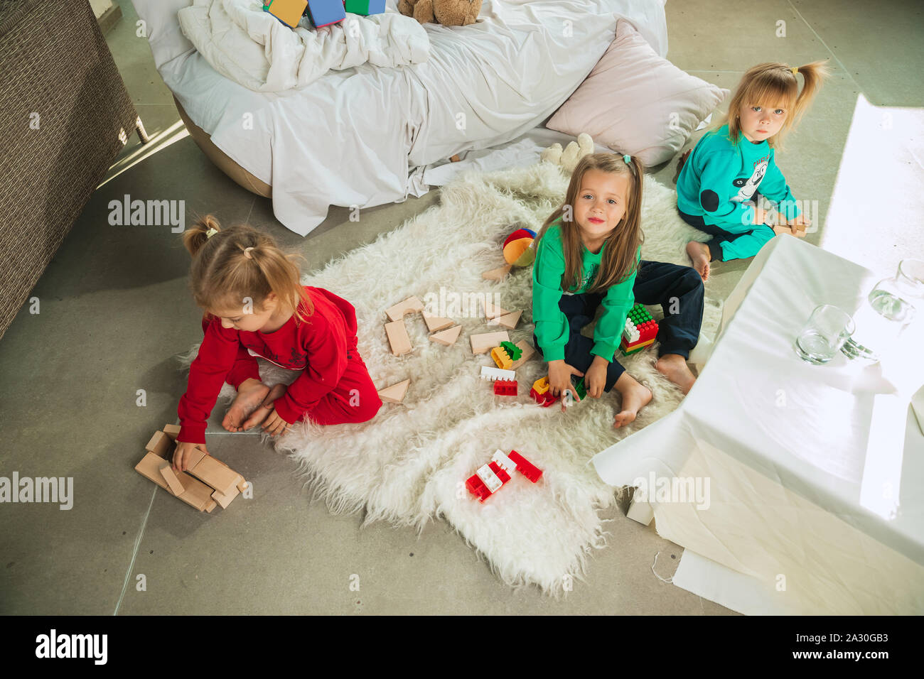 Little girls in soft warm pajamas playing at home. Caucasian children in colorful clothes having fun together. Childhood, home comfort, happiness. Sitting on the floor and making constructor. Stock Photo