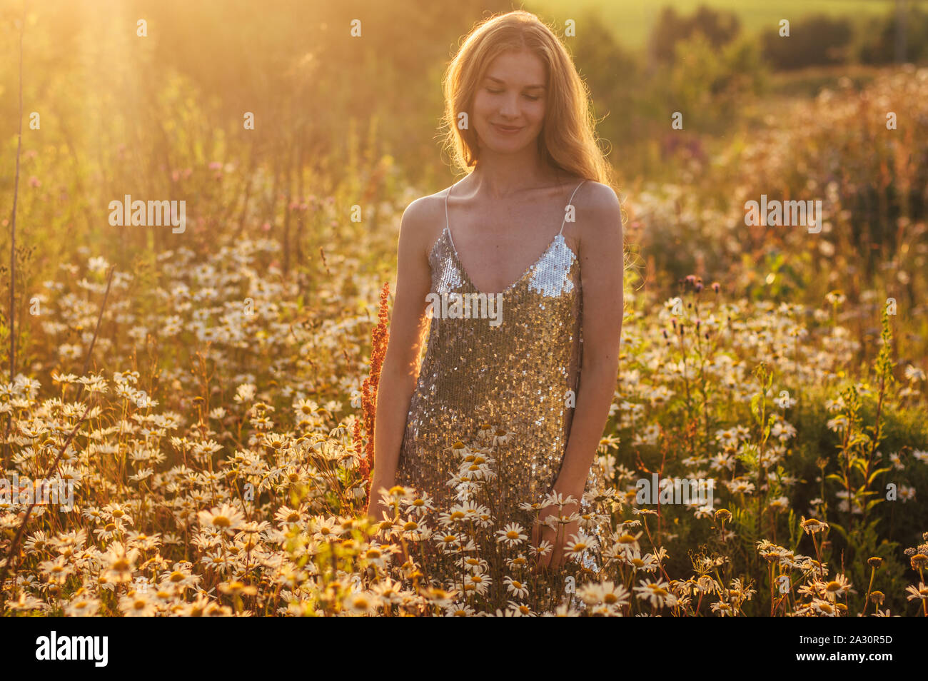 Smiling girl with closed eyes in blinking dress staying among chamomile ...