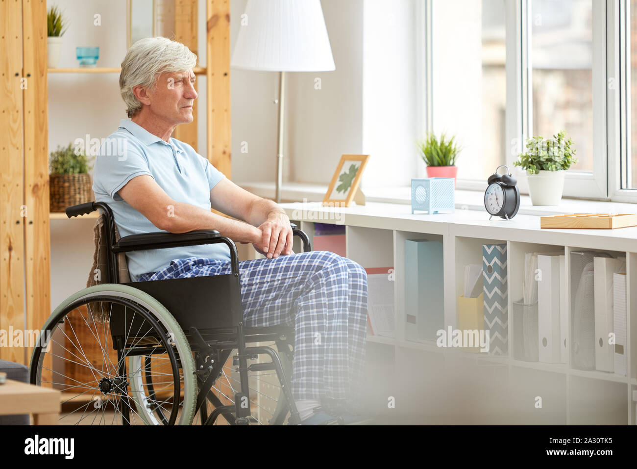 Senior patient sitting in wheelchair and thinking about something while looking out the window at nursing house Stock Photo