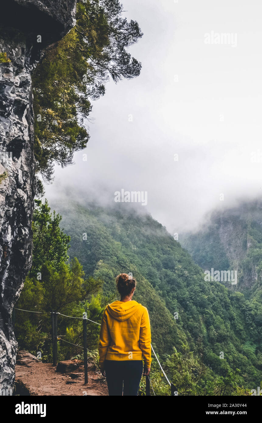 Young woman in yellow sweatshirt on viewpoint in Levada Caldeirao Verde, Madeira, Portugal. Green scenic mountains in mist, foggy landscape. Female traveler. Instagram, hipster filter. Vertical photo. Stock Photo