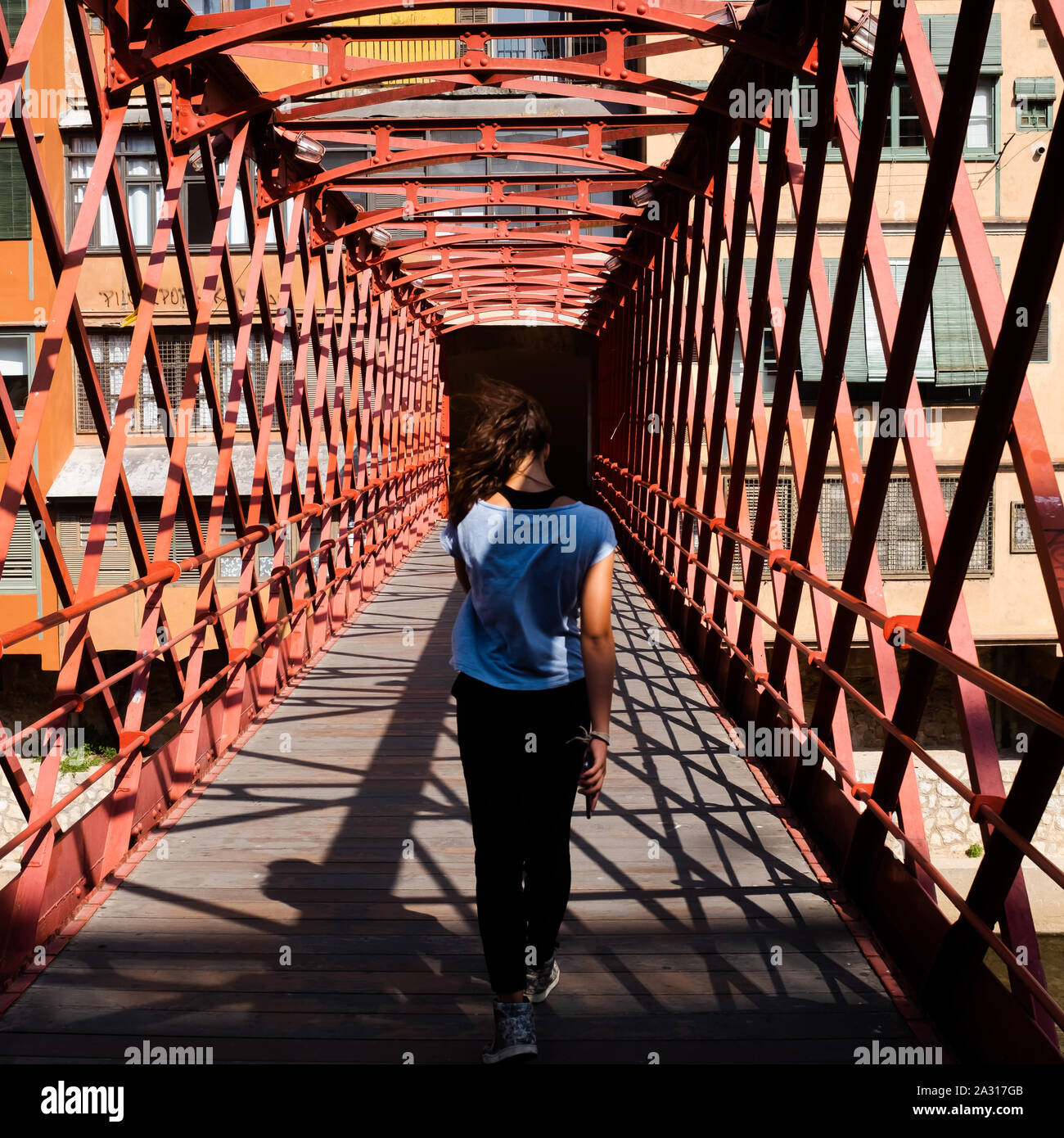 Back view of a young girl with fluttering brown hair, wearing a blue summer t-shirt and crossing the Pont de les Peixateries Velles in Gerona, Spain. Stock Photo