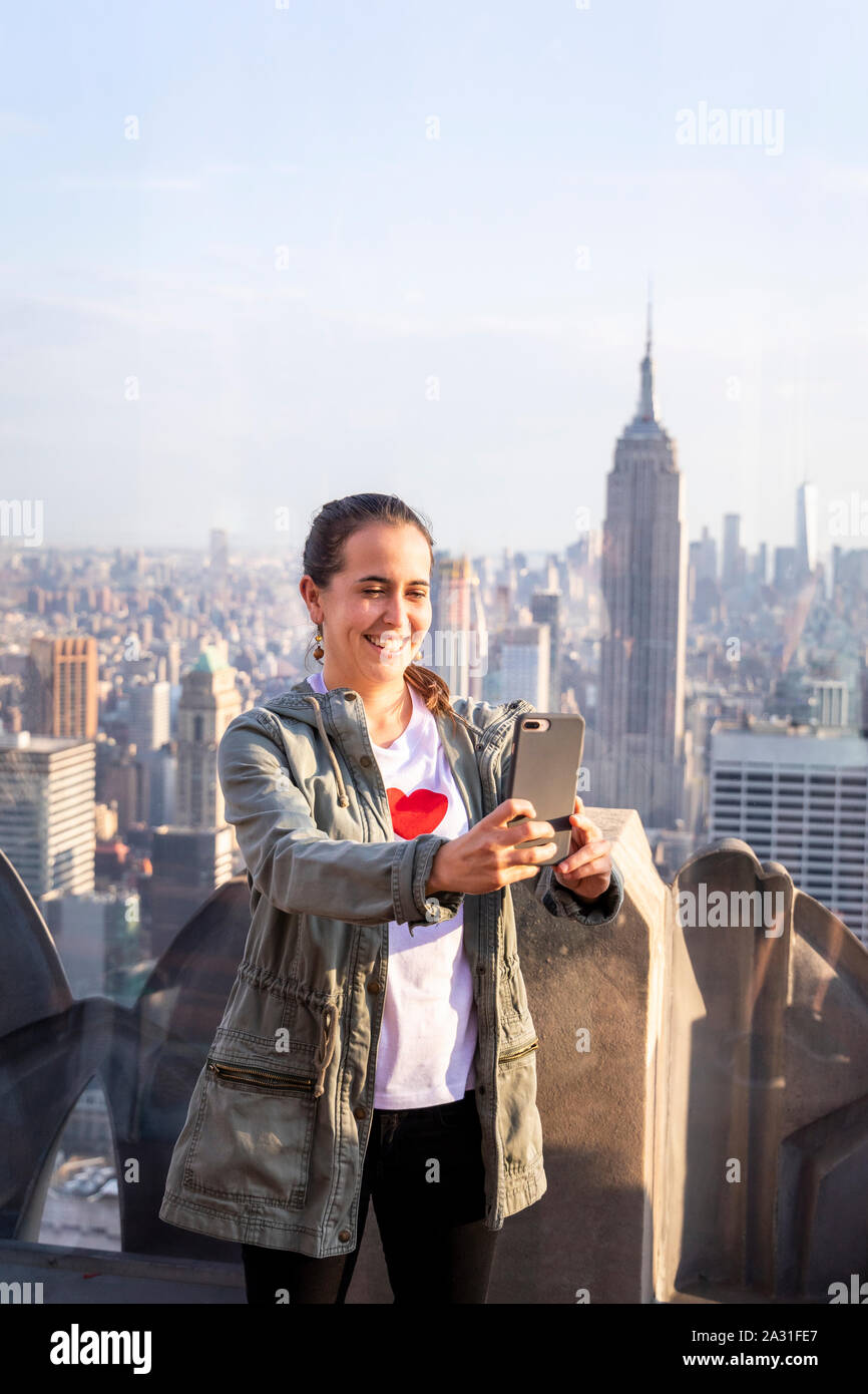 Female tourist takes a selfie on Top of the Rock Observatory with the Empire State Building in the distance, New York City, USA. Stock Photo