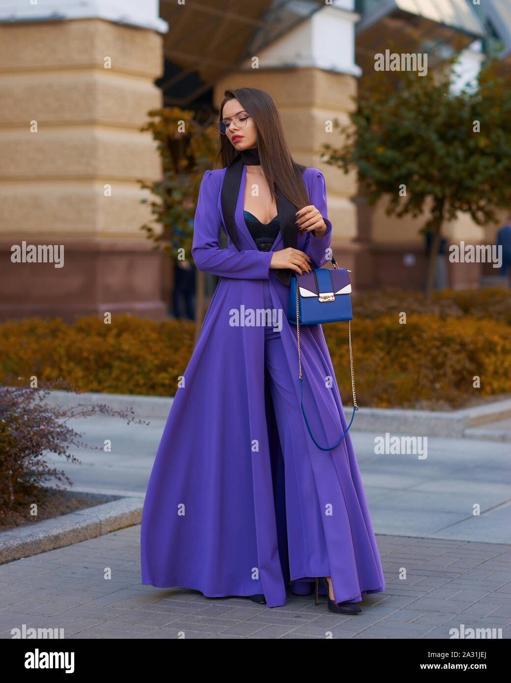 Young beautiful caucasian woman with long straight hair and makeup in purple trousers and long coat cape standing and posing at city street in autumn Stock Photo