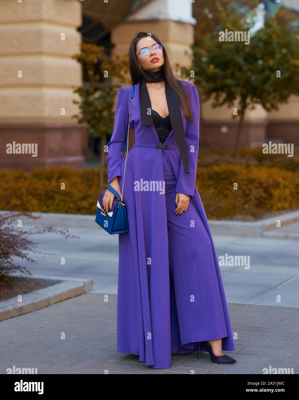 Young beautiful caucasian woman with long straight hair and makeup in purple trousers and long coat cape standing and posing at city street in autumn Stock Photo