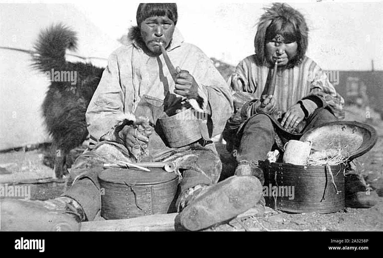 Eskimo couple smoking pipes seated beside carved wooden boxes and tools ...