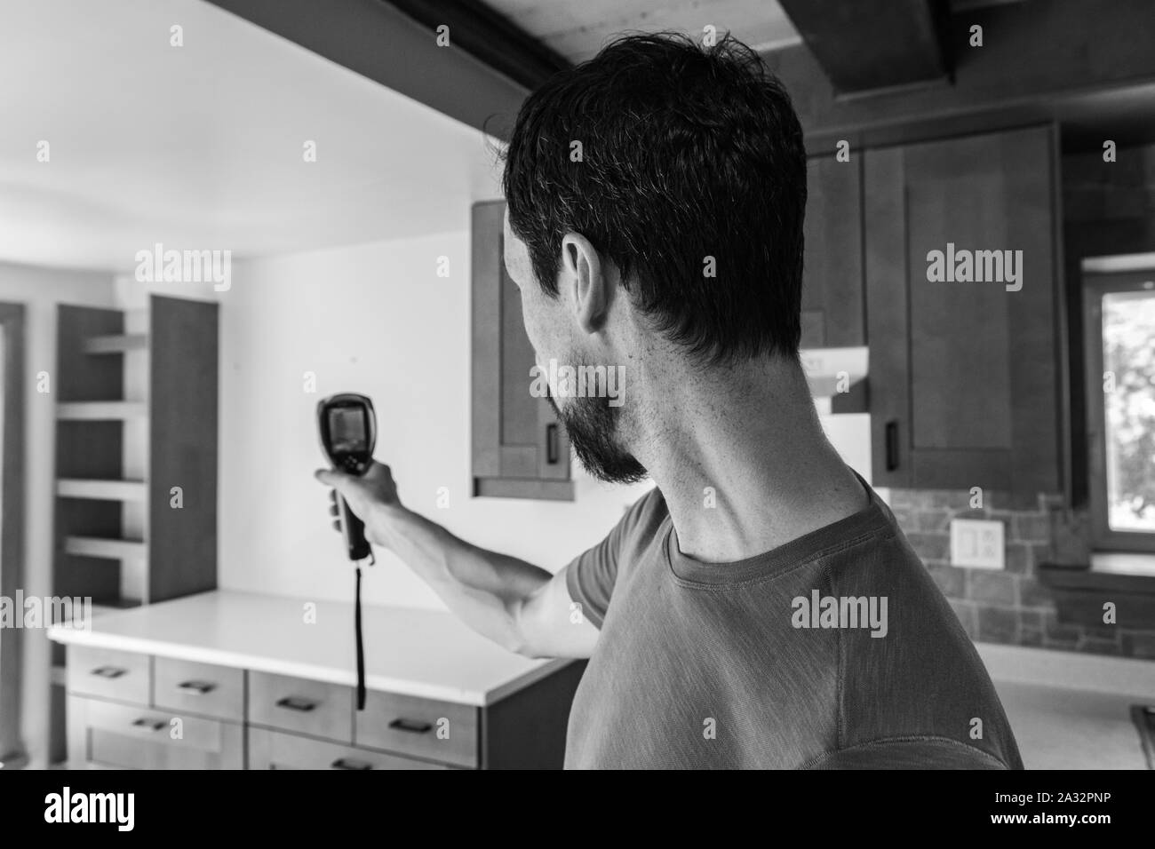 A close up and monochrome view of a man using a small hand held thermal imagery device inside a family kitchen during a building survey, with copy-space. Stock Photo