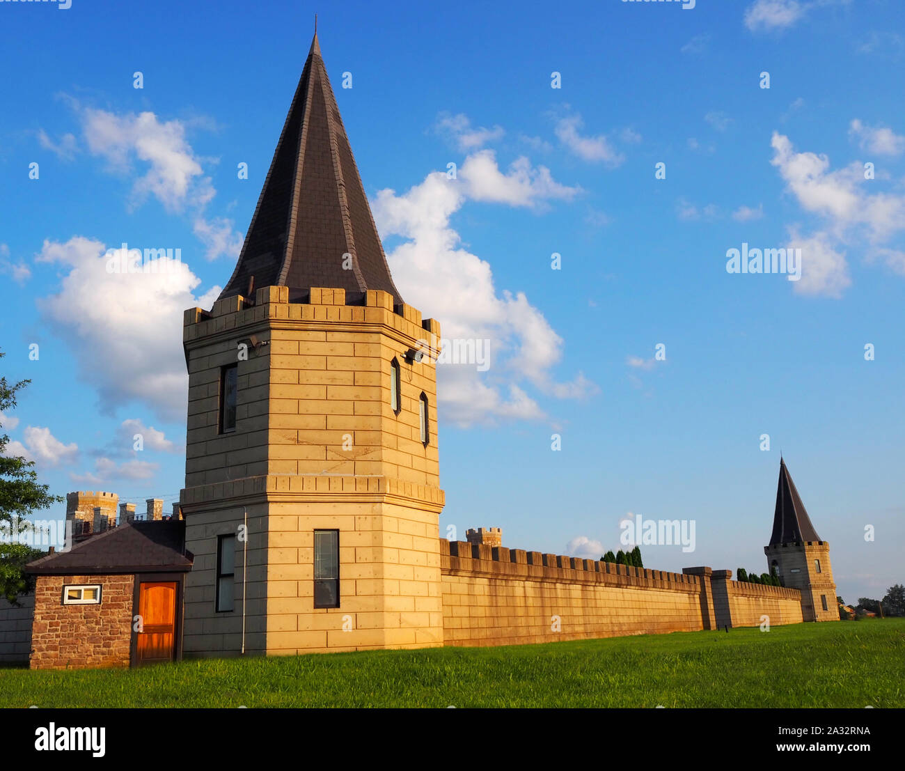 A turret marks the corner of large castle sprawling into the background surrounded by green grass and a vibrant blue sky with wispy clouds. Stock Photo