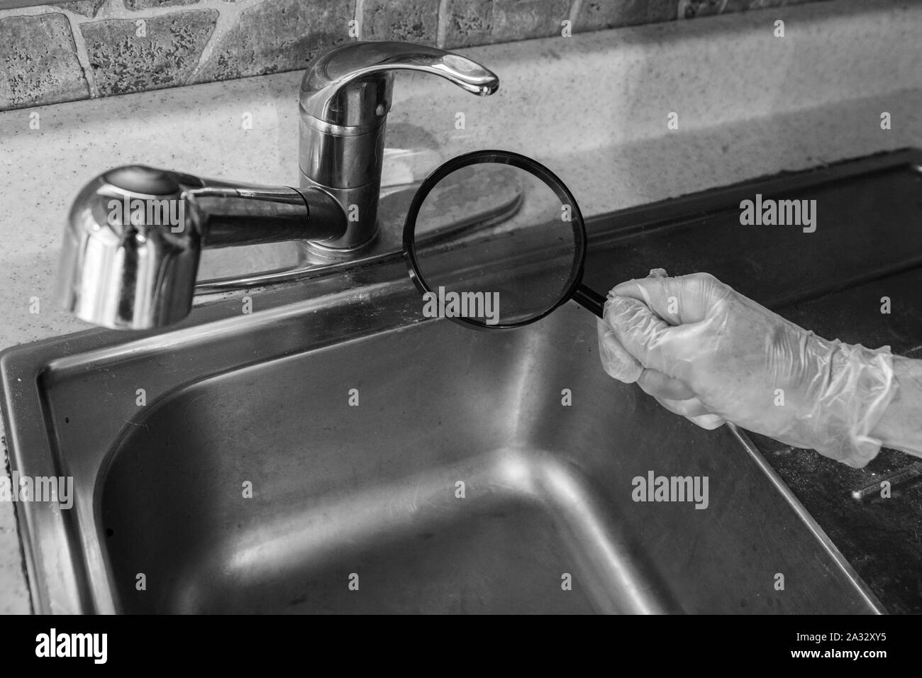 A close up and monochrome view of a person inspecting a kitchen sink and faucet with a magnification glass, checking for imperfections with copy space. Stock Photo